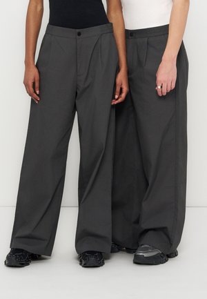 Dwie osoby w luźnych, ciemnoszarych spodniach i czarnych trampkach stojące obok siebie na białym tle.