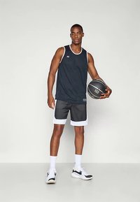 Mlad moški v črnem košarkarskem dresu in hlačah drži črno košarko, nosi bele nogavice in črno-bele superge, stoji na enobarvni podlagi.