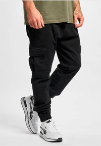 Zwarte cargo broek met zijzakken, elastische boorden en een losse pasvorm, gecombineerd met wit-grijze sneakers met een zwarte accentlijn.