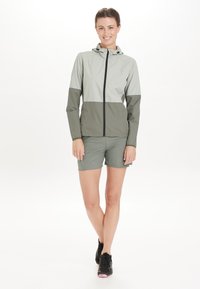 Lichte, twee-kleurige jas in grijs en olijfgroen, met een rits aan de voorkant, een capuchon en elastische manchetten. Gecombineerd met bijpassende shorts en zwarte sneakers.