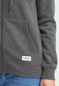 Felpa grigia con zip realizzata in tessuto morbido, dotata di una tasca frontale, polsini elasticizzati e una piccola etichetta bianca con il logo del marchio.