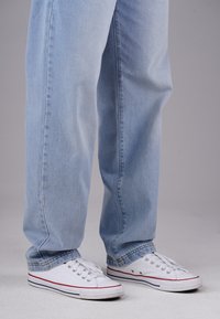 Person, der hellblaue gerade geschnittene Jeans und weiße flache Canvas-Sneaker trägt, steht auf einem hellgrauen Boden.