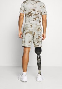 Pantalones cortos de camuflaje gris claro para hombre y camisa a juego, hechos de una tela suave y transpirable. Pierna protésica negra visible, zapatillas blancas.