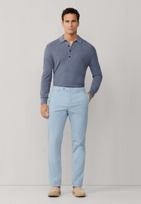 Homme debout portant un polo à manches longues bleu, un pantalon bleu clair et des chaussures beige à enfiler, sur un fond uni.