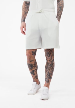 Homme portant un short blanc et des baskets, montrant des jambes et des bras tatoués avec des motifs noirs détaillés, y compris des roses, des papillons et du texte.