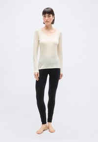 Γυναίκα που στέκεται ξυπόλυτη φορώντας μια κρεμ μακρυμάνικη μπλούζα και μαύρα κολάν μπροστά σε ένα απλό φωτεινό φόντο.