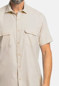 Camisa de manga corta en un color beige claro, confeccionada con una tela texturizada que presenta dos bolsillos en el pecho y cierres de botón.