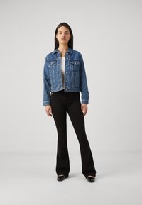 Giacca di jeans blu con chiusure a bottoni, due tasche sul petto e un design cropped, abbinata a pantaloni neri a campana e scarpe nere.