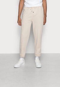 Beige sweatpants with a drawstring waist, tapered cuffs, and side seams, paired with white sneakers and visible white socks.