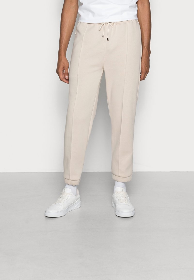 Beige sweatpants with a drawstring waist, tapered cuffs, and side seams, paired with white sneakers and visible white socks.