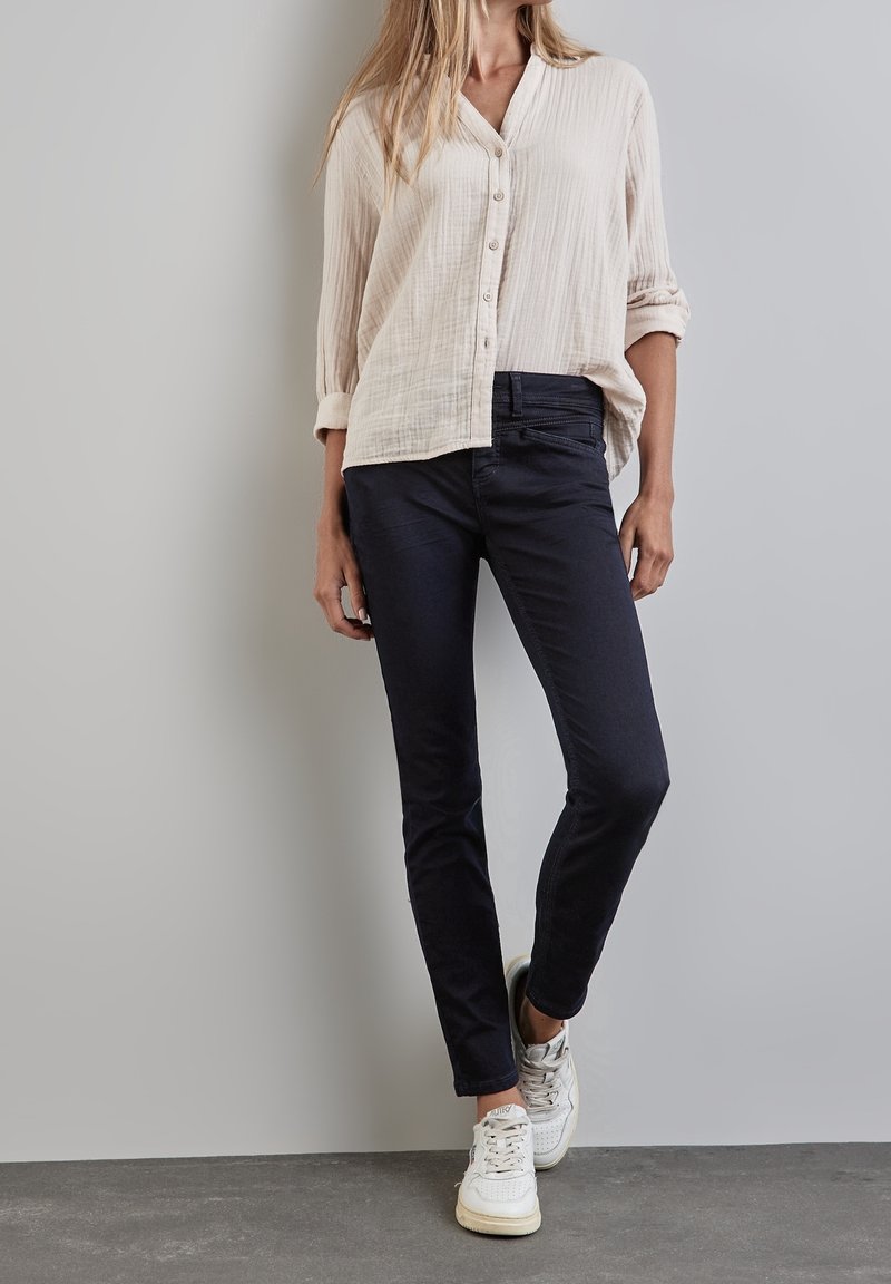Femme portant une chemise beige clair à boutons avec les manches retroussées, un pantalon slim foncé et des baskets blanches, se tenant devant un mur gris uni.
