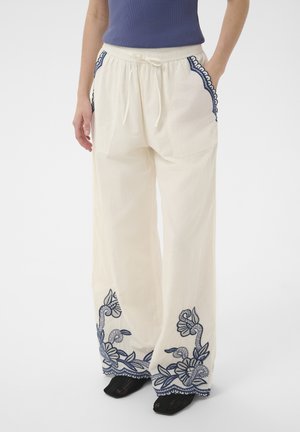 Pantalon blanc ample avec broderie florale bleu marine sur les poches et les ourlets, porté avec des chaussures noires et un haut sans manches bleu marine.