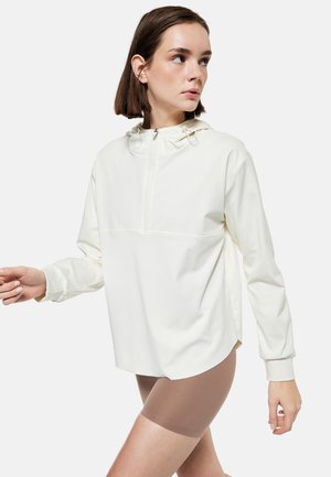 Jonge vrouw draagt een oversized witte hoodie met een rits aan de voorkant en beige fietsbroekjes, kijkt naar links.