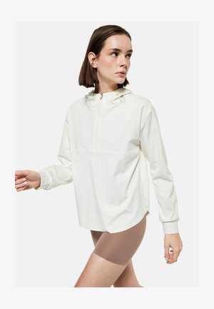 Jeune femme portant un pull à capuche blanc oversize avec une fermeture éclair à l'avant et un short de cyclisme beige, regardant vers sa gauche.