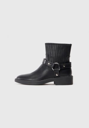 Stivaletto in pelle nera con tomaia in tessuto a costine, una larga cinghia con hardware argentato e un tacco basso a blocco.