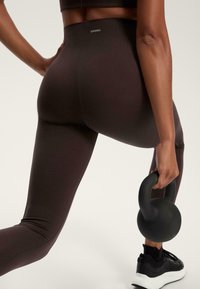 Schwarze Leggings mit glatter Textur und hohem Bund, mit einem kleinen Logo auf der Rückseite. Das Model hält eine schwarze Kettlebell.
