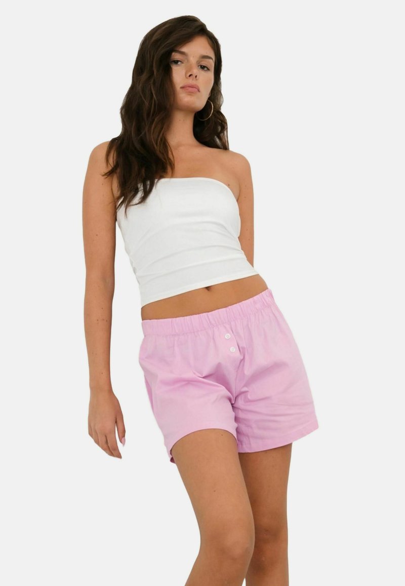Jeune femme aux longs cheveux foncés portant un haut blanc sans bretelles et un short rose pâle à taille élastique, debout devant un fond blanc.