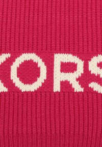 Tissu tricoté rose avec de grandes lettres blanches formant le mot "KORS". La texture semble douce et côtelée avec un design cohérent à travers tout le tissu.