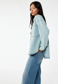 Camicia in denim azzurro chiaro con una vestibilità rilassata, maniche lunghe e spacchi laterali. Presenta una tasca sul petto e dettagli di cucitura minimal. Abbinata a jeans blu.