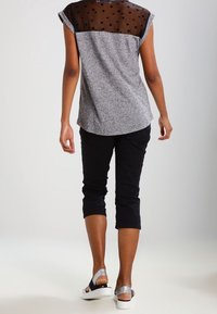 Nainen, jolla on päällään harmaa paita mustalla läpikuultavalla pistekuviolla, mustat lyhyet housut ja valkoiset korkeat sandaalit, seisoo selin.