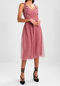 Vaaleanpunainen polven mittainen mekko, jossa on kukka-applikaatioita, poimutettu rintamuksen osa ja läpinäkyvä hame. Yhdistetty mustiin hihnallisiin korkokenkiin.