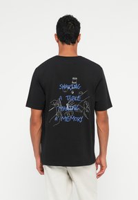 Vīrietis ģērbjas melnā T-kreklā ar zilu tekstu "DALOT GALDU, VEIDOJOT ATMIŅAS" un ar līniju zīmējumiem, kas attēlo rokas un priekšmetus aizmugurē.