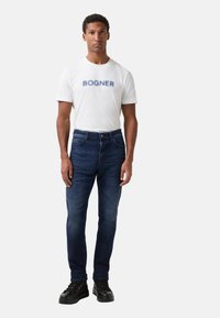 Weißes Baumwoll-T-Shirt mit "BOGNER" Druck, kombiniert mit dunkelblauen Jeans. Schwarze Schnürschuhe runden das Outfit ab.