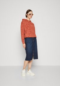 Sudadera con capucha de color óxido con textura estampada, combinada con una falda lápiz de denim oscuro con una abertura frontal, y zapatillas blancas.