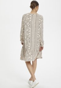 Långärmad beige klänning med svarta geometriska mönster, rundad halsringning och volang vid nederkanten, bärs med vita platåskor.