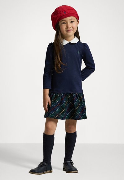 Tengerészkék hosszúujjú felső fehér gallérral, zöld, kék és piros mintás kockás szoknyával, fekete térdharisnyával és fekete cipővel párosítva. Piros kötött baret.