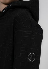 Felpa nera con cuciture blu, caratterizzata da un motivo testurizzato e una patch del logo sulla manica. Il materiale sembra morbido ed elastico.