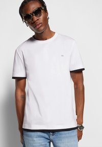 T-shirt bianco in cotone con bordi neri alle maniche e all'orlo, con un piccolo logo "CK" sul petto. Indossato con jeans azzurri chiari e occhiali da sole.