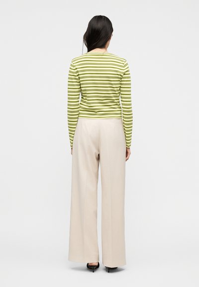 Γυναίκα με μακριά σκούρα μαλλιά που φοράει μακρυμάνικη μπλούζα με πράσινες και λευκές ρίγες και μπεζ παντελόνι φαρδύ στα πόδια, γυρισμένη προς τα πίσω σε λευκό φόντο.