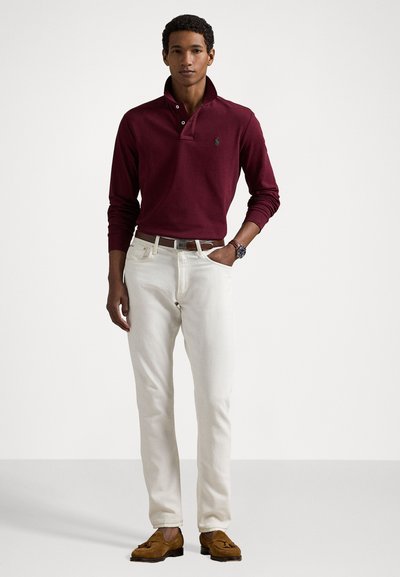 Burgundska polo majica s dolgimi rokavi in ovratnikom, bele jeans hlače, rjavi mokasini, ura na zapestju in roke v žepih.