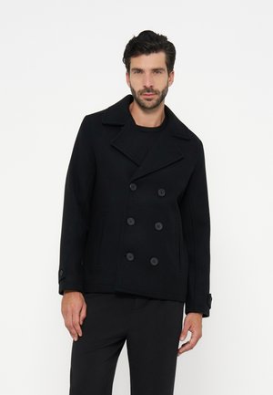 Homme portant un caban noir croisé et un pantalon noir, debout devant un fond clair uni.