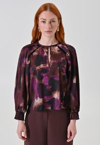 Blusa a maniche lunghe in viola scuro e marrone, con apertura a chiave sul davanti, spalle plissettate e polsini arricciati. Tessuto liscio e fluido.