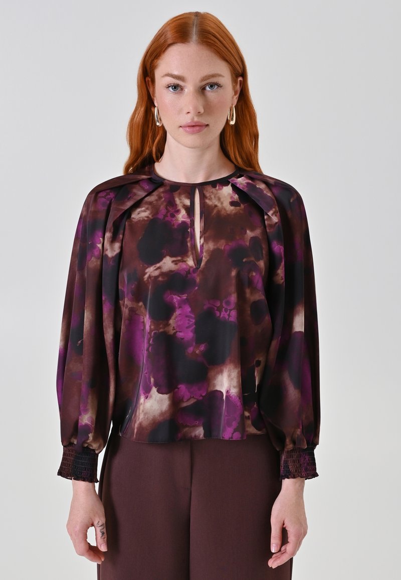 Blusa a maniche lunghe in viola scuro e marrone, con apertura a chiave sul davanti, spalle plissettate e polsini arricciati. Tessuto liscio e fluido.
