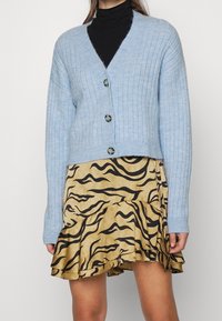 Vaaleansininen resorineuleinen neuletakki V-kauluksella ja kolmella napilla, yhdistettynä keltaiseen hameeseen, jossa on musta tiikerinraidallinen kuvio.