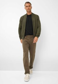 Olijfgroene ritscardigan over een zwart shirt, gecombineerd met een paarse broek en beige sneakers. Eenvoudige, ribbelige textuur op de cardigan.