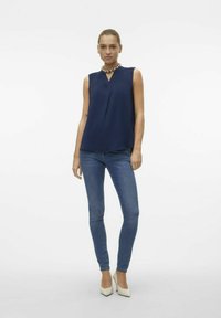 Blouse sans manches bleu marine avec un décolleté en V, assortie à un jean skinny bleu et des chaussures à talons blancs. Tissu lisse et coupe décontractée.
