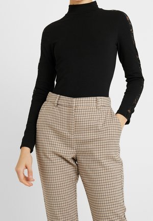 Kobieta w czarnym, długim rękawie golfie z koronkowymi detalami na rękawach oraz beżowych, wysokim stanem, w kratkę spodniach z kieszeniami.