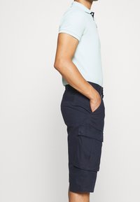 Polo shirt bleu clair avec un col porté avec un short cargo navy comportant des poches latérales et un devant plat, fabriqué dans un tissu doux.