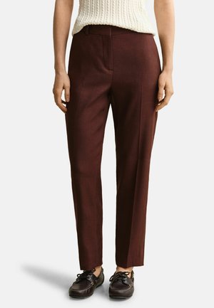 Femme portant un pantalon marron foncé taille haute, droit, avec des mocassins noirs à lacets et un haut sans manches crème en maille torsadée.