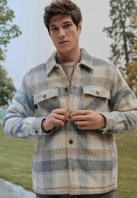 Kockovaná vlnená bunda v šedej, béžovej a bielej farbe s dvoma náprsníkmi, gombíkovým zapínaním a golierom. Model stojí vonku, s rukami na bunde.