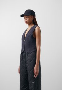 Džínová vesta v tmavě modré barvě s hlubokým výstřihem do V, třemi knoflíky a bez rukávů, spárovaná s prošívanými černými kalhotami s elastickým pasem.