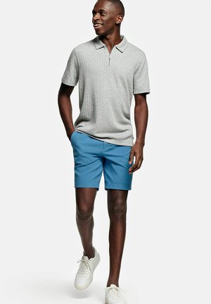 Jonge man glimlacht, loopt met handen in de zakken, draagt een lichtgrijze korte mouw polo, blauwe korte broek en witte sneakers.