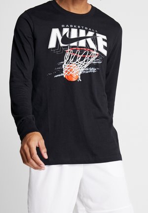 Homme portant un t-shirt noir à manches longues avec un graphique de panier de basket et de ballon ainsi qu'un logo Nike blanc, associé à un short blanc.