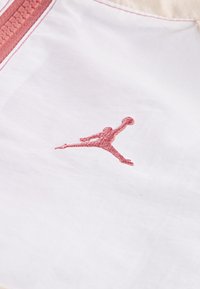 Balta auduma virsma ar gludu struktūru, rotāta ar sarkanu izšūtu Jumpman logo. Iekļauts sarkans rāvējslēdzis ar teksturētu vilkšanas cilni.
