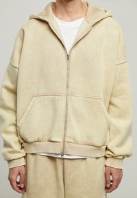 Felpa beige con zip realizzata in tessuto morbido, dotata di tasca a marsupio, polsini a coste e cappuccio foderato per un comfort aggiuntivo. Lavabile in lavatrice.