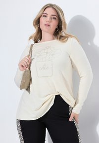 Ecrufarbene Langarmbluse mit floralem Design aus Perlen; kombiniert mit schwarzen Hosen mit Leopardenmuster-Akzenten an den Seiten. Enthält eine kleine Handtasche.